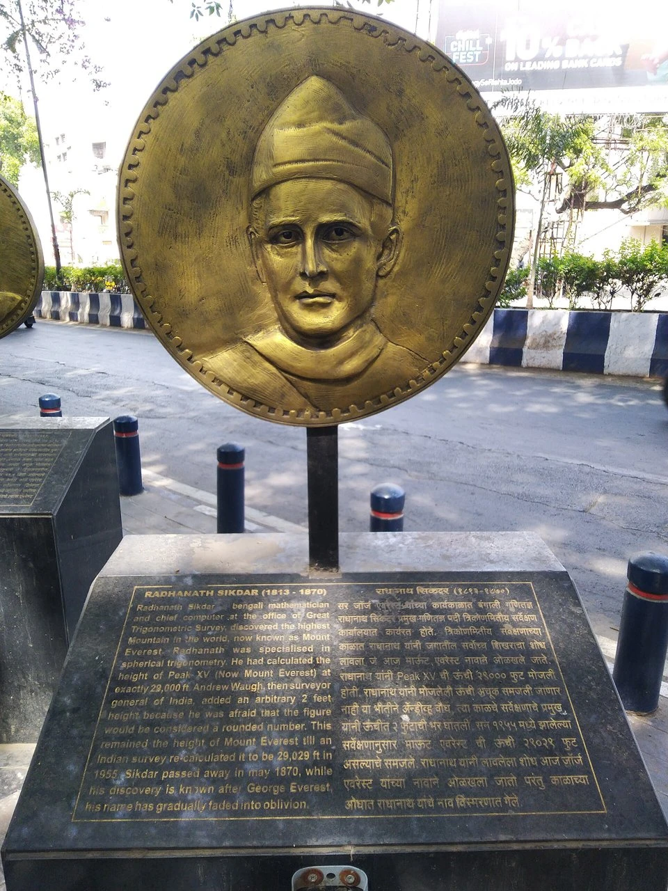 A bronze medallion of Radhanath Sikdar on a pedestal beside a Pune street, above a stone plaque with text in English and Marathi.