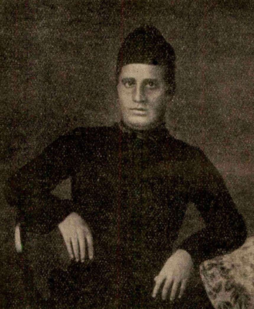 Portrait of Radhanath Sikdar, seated, in a dark buttoned coat and a dark cap, one hand resting on each knee.