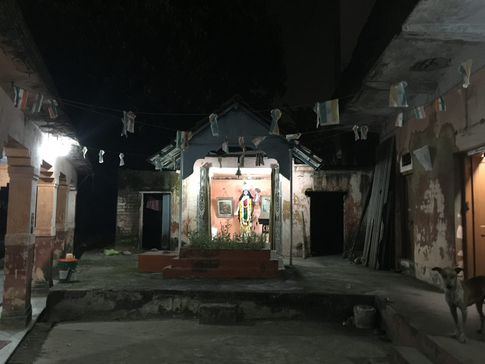 The courtyard of the Hindu Mission at night, strung with faded paper Indian flags, with a lit shrine to a blue-skinned Vishnu at the far end