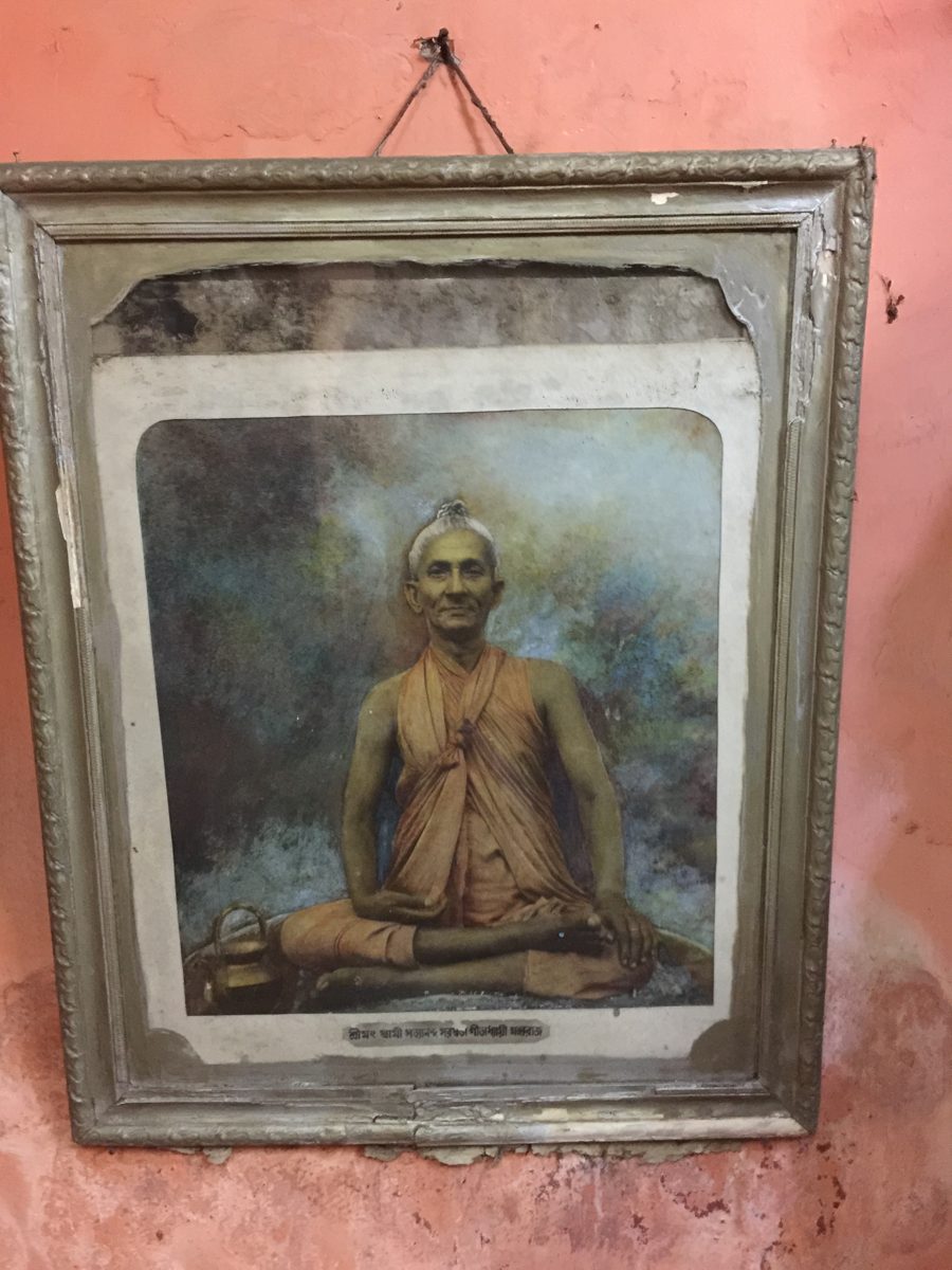 A framed, hand-tinted photograph of an elderly monk in saffron robes seated cross-legged, hanging on a pink wall