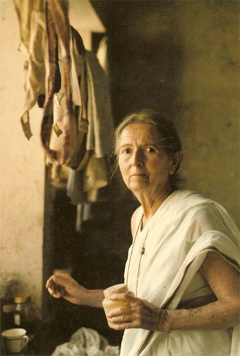 Savitri Devi as an old woman in a white sari, holding a cup, in a dim room